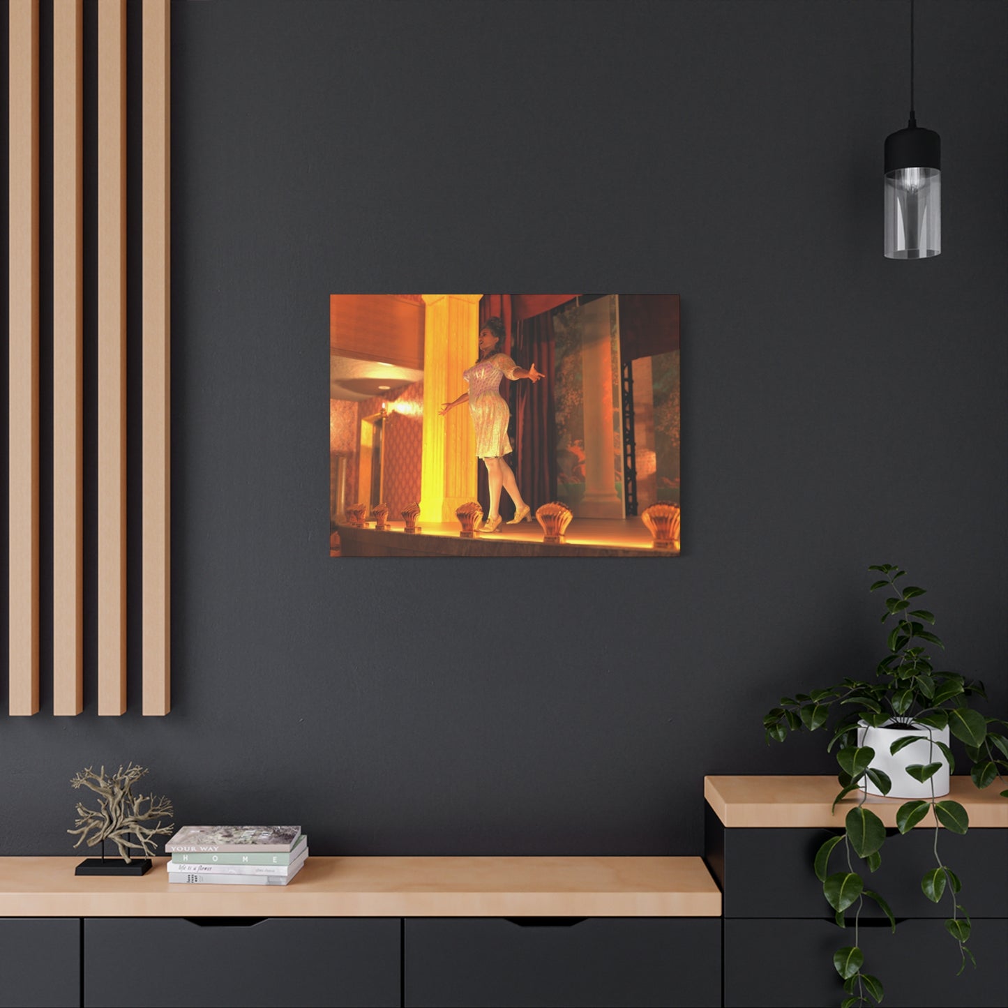 A rectangular wall art print showing a warmly lit figure in a white dress, displayed on a dark charcoal wall above a light wood console with books, a small plant, and decor.