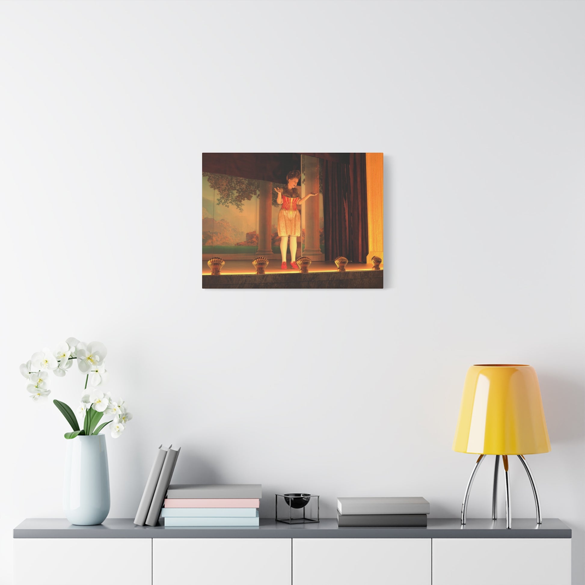 A framed wall art print depicting a classical scene with a figure in a white dress standing in a doorway under warm golden light, displayed on a white wall above a console table with a yellow lamp, books, and a vase of white flowers.