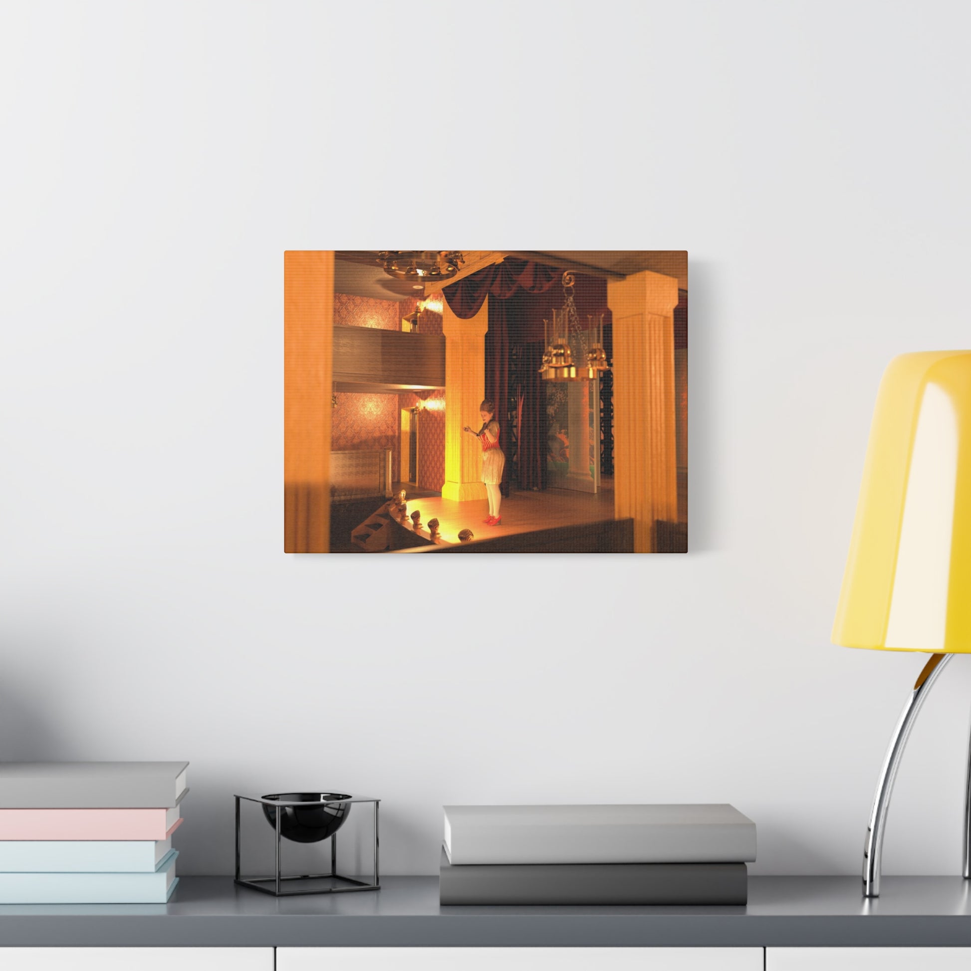 A framed wall art print depicting a warmly lit stage scene hangs on a white wall above a gray console table styled with books, a small black bowl stand, and a yellow lamp.