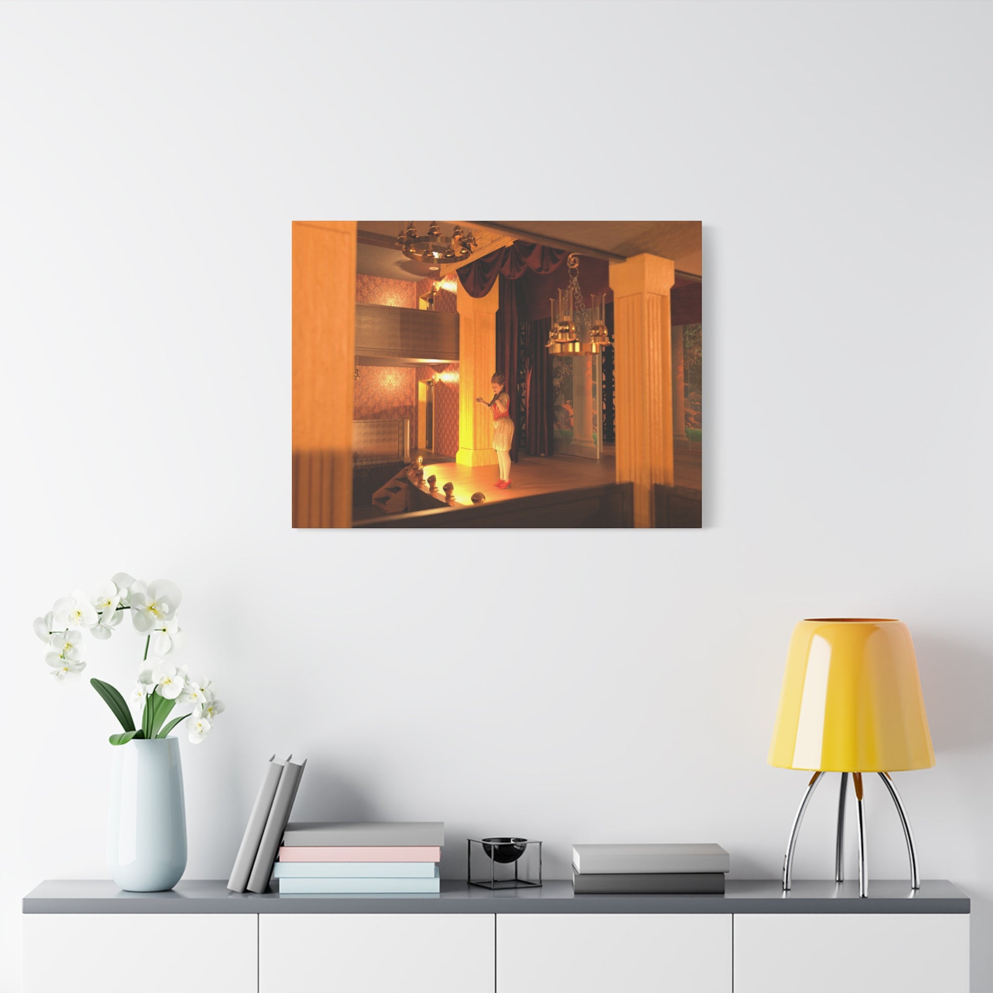 A rectangular wall art print showing a warmly lit interior scene with orange and yellow tones, displayed above a console table with a yellow lamp, books, and white flowers in a bright room.