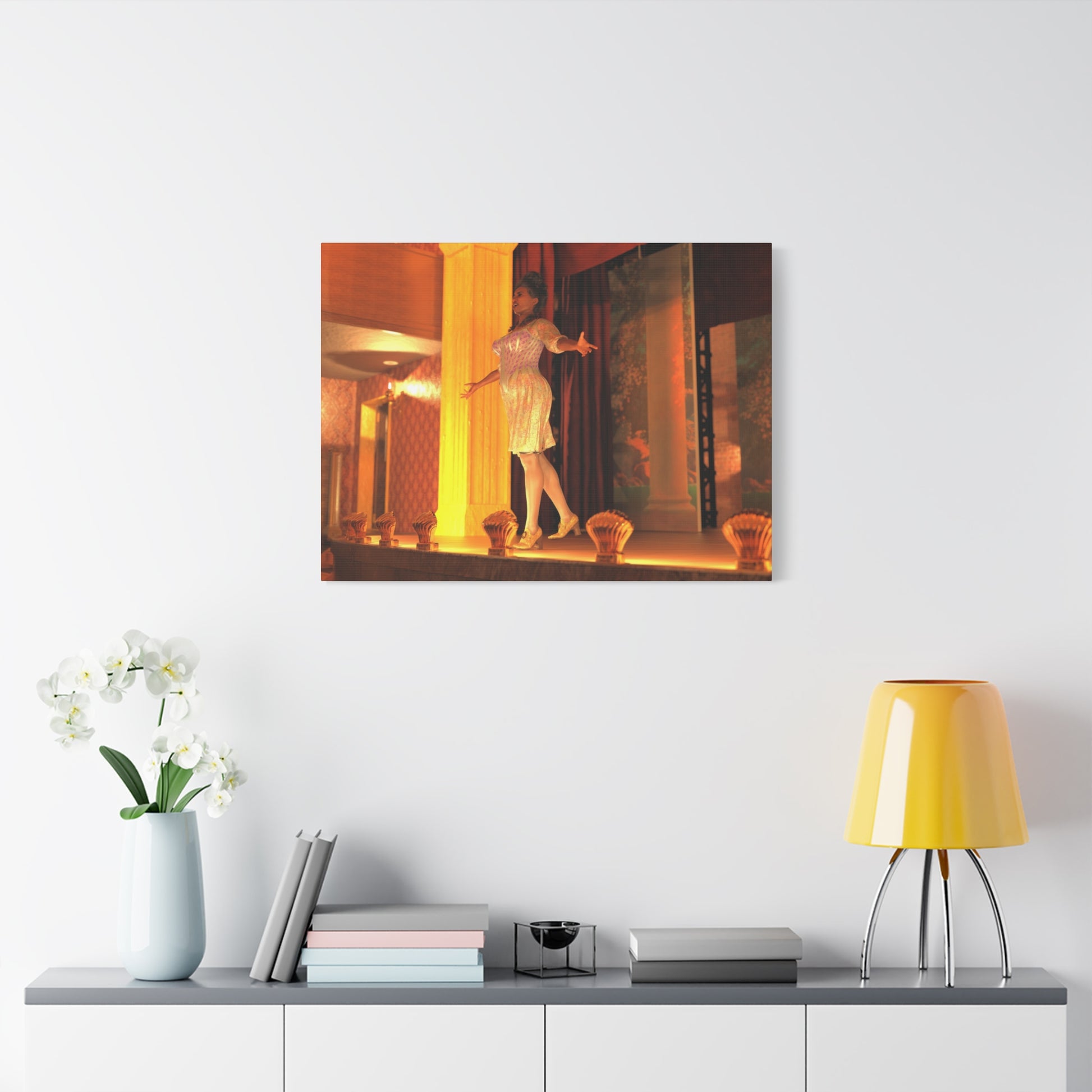 A framed wall art print depicting a woman in a white dress dancing in a warmly lit interior, displayed on a white wall above a console table with a yellow lamp, books, and a vase of white flowers.