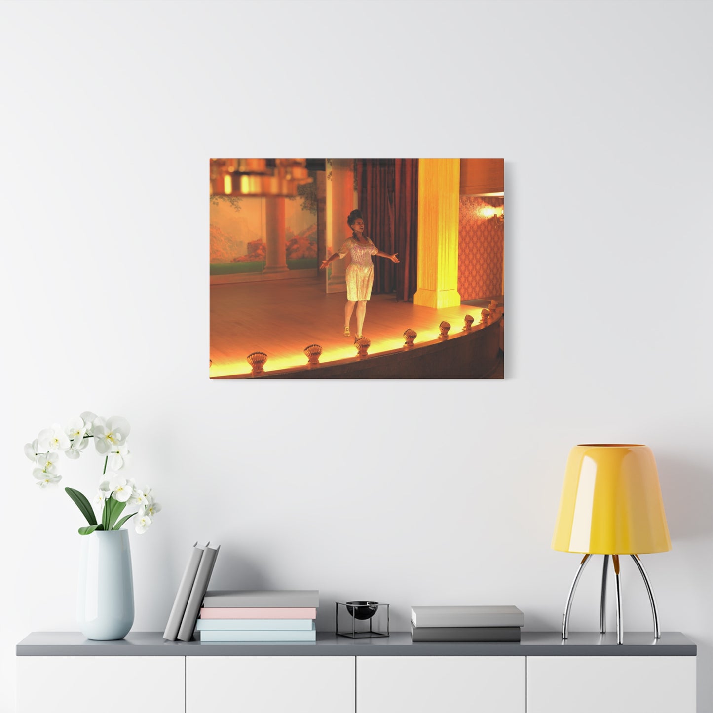 A framed wall art print depicting a dancer on a warmly lit stage, displayed above a console table with books, a vase of white flowers, and a yellow lamp in a modern room setting.