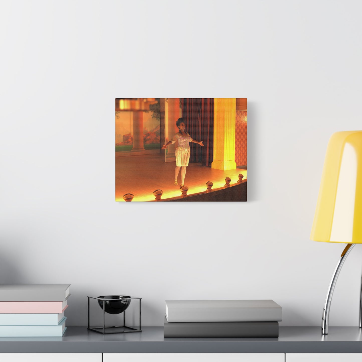 A rectangular wall art print showing a woman dancing on a warmly lit stage, displayed on a white wall above a shelf with books, a small black object, and a yellow lamp.
