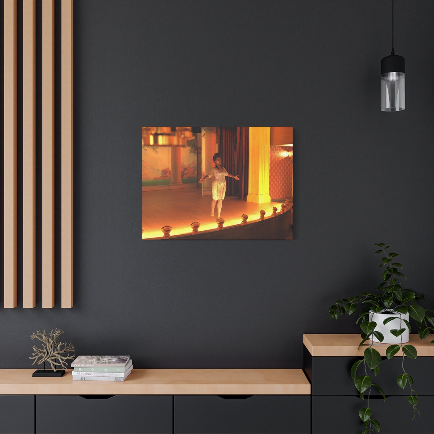 A rectangular wall art print showing a warmly lit stage scene with a person, mounted on a dark charcoal wall above a light wood console with decor and a hanging pendant lamp.