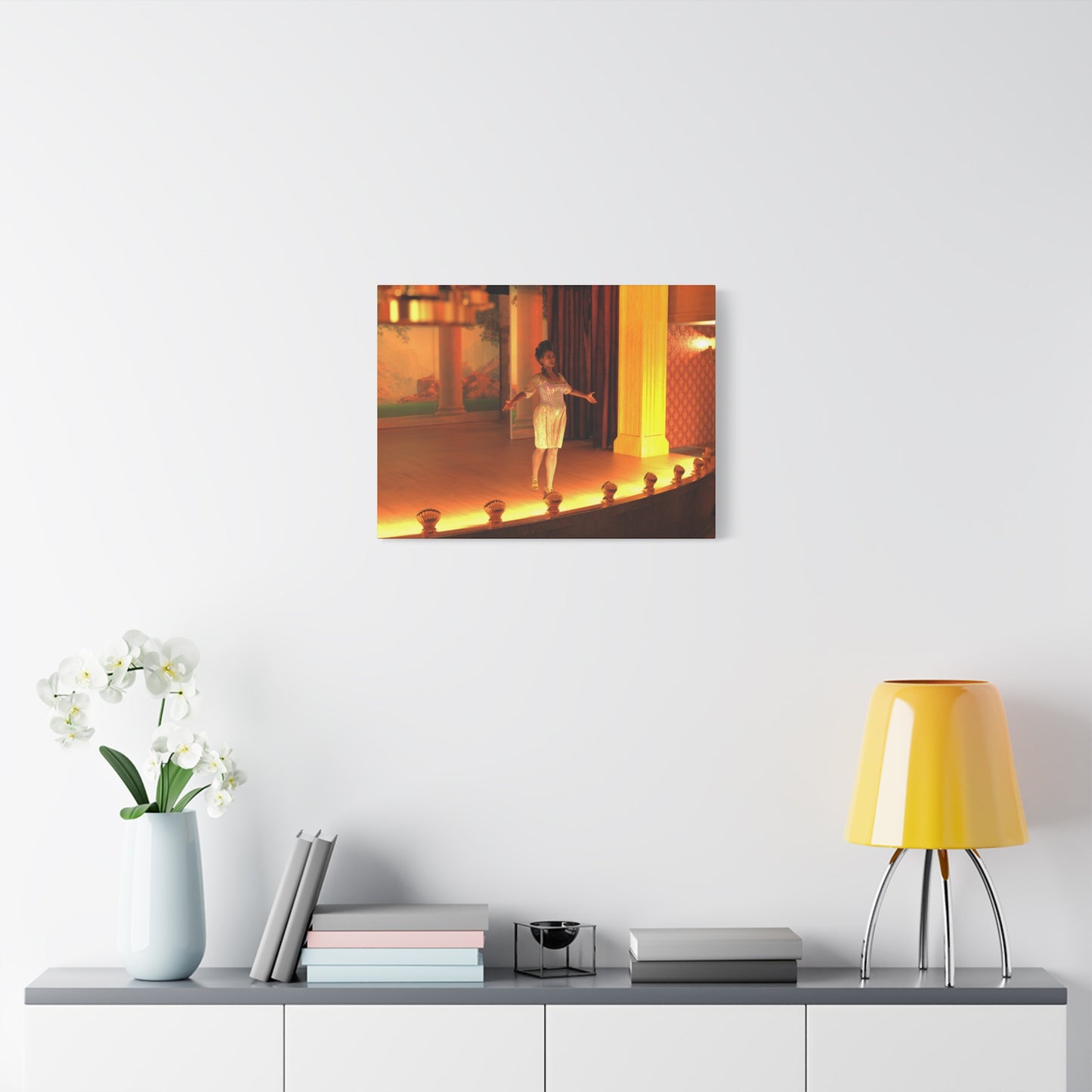 A framed wall art print showing a child dancing on a warmly lit stage, displayed above a console table with a yellow lamp, books, and a vase of white flowers in a bright modern room.