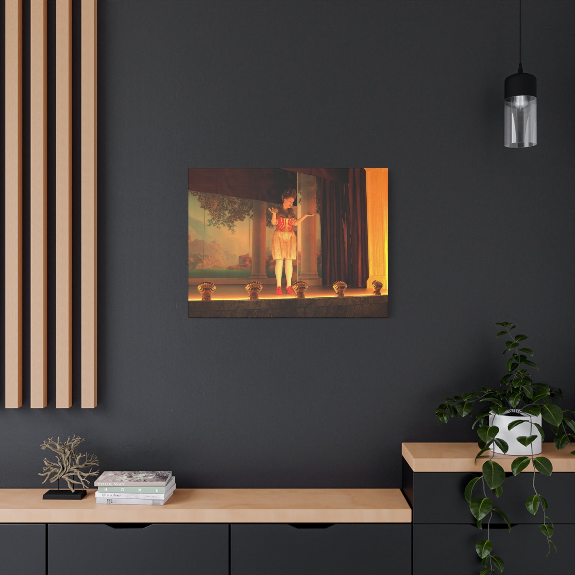 A framed art print depicting a classical scene with a figure in a red dress standing in a doorway, displayed on a dark gray wall in a modern room setting with a wooden console, plants, and a hanging light.