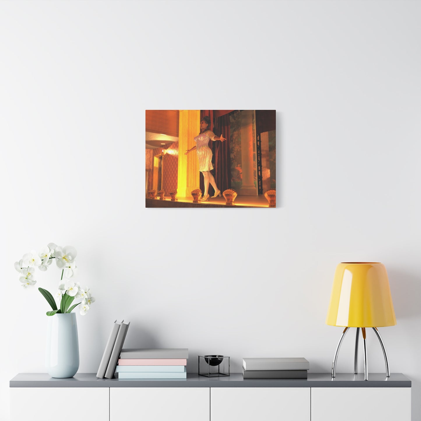 A framed wall art print depicting a woman in a white dress dancing in warm orange and yellow light, displayed on a white wall above a console table with a yellow lamp, books, and a vase of white flowers.