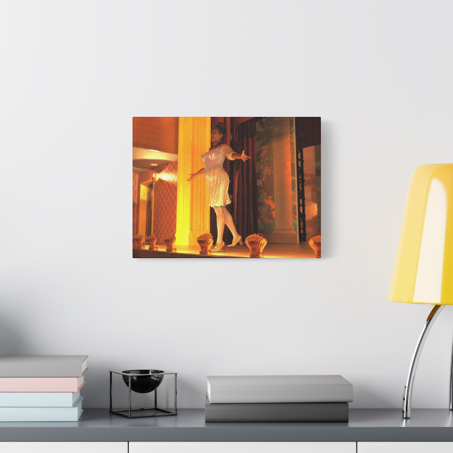 A framed wall art print depicting a woman in a white dress in a warmly lit interior, displayed on a light gray wall above a shelf with books and a yellow lamp.
